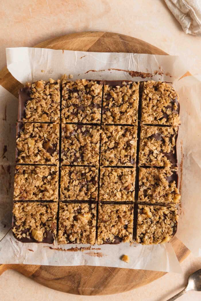 Overhead view of a pan of oatmeal chocolate bars cut into 16 small squares. The bars are sitting on a piece of parchment paper on top of a round cutting board.