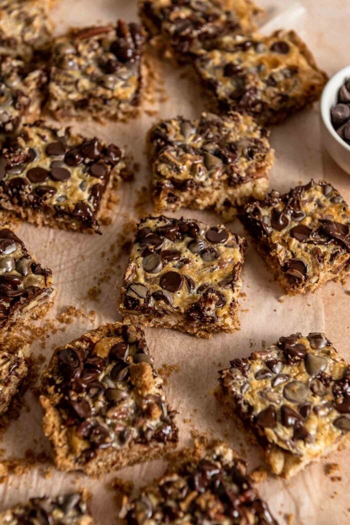 A number of vegan magic cookies bars scattered around a piece of parchment paper. The bars have chocolate chips, pecans and coconut in them.