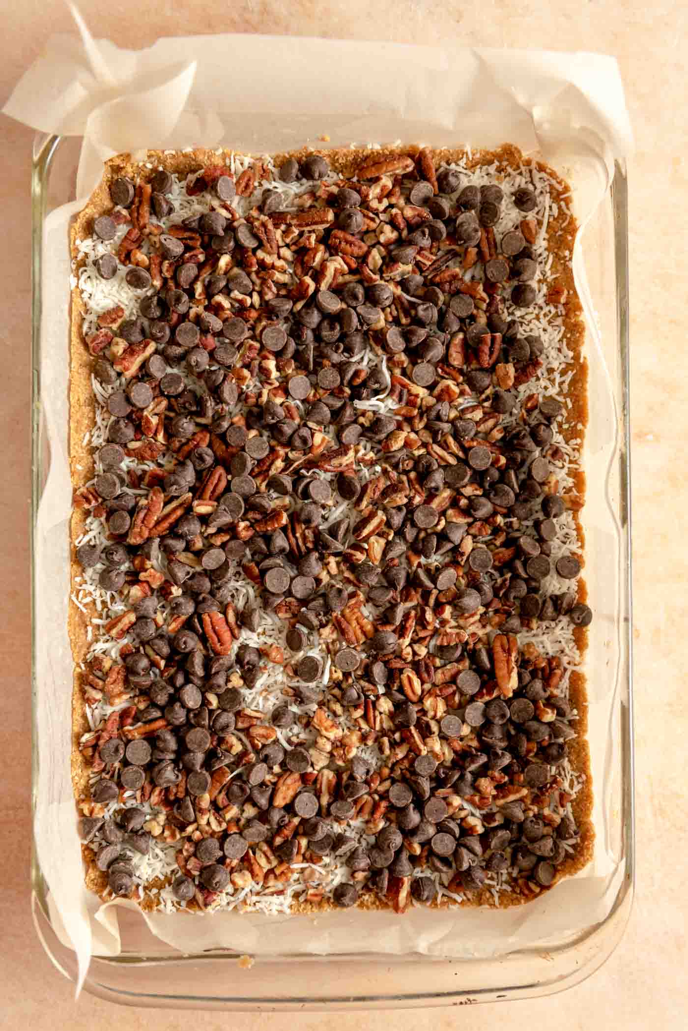 Chocolate chips, pecans and shredded coconut sprinkled over magic cookie bars in a 9x13-inch glass backing dish lined with parchment paper.
