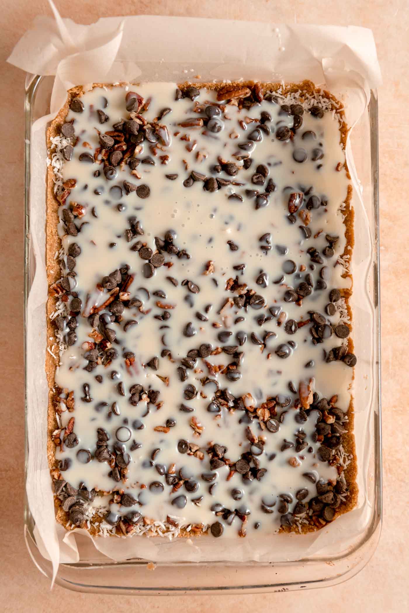 Coconut condensed milk poured into a 9x13-inch glass pan of seven layer magic cookie bars before they've been baked.