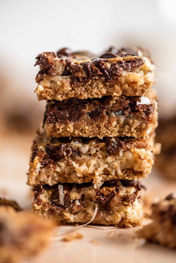 A stack of 4 seven layer cookies bars with a layer of graham cracker crust and a gooey layer of coconut, chocolate chips and pecans.