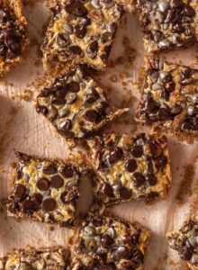 Overhead view of a batch of 7 layer cookies bars scattered on a counter. The bars are topped with chocolate chips, coconut and pecans.