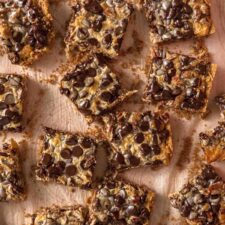 Overhead view of a batch of 7 layer cookies bars scattered on a counter. The bars are topped with chocolate chips, coconut and pecans.