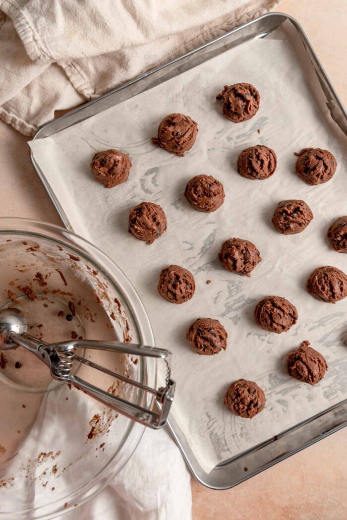 A cookie scoop resting in a glass mixing bowl beside a sheet pan of chocolate snowball cookies.