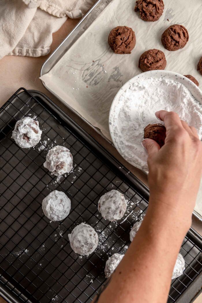 A hand dipping a chocolate snowball cookie in a bowl of icing sugar and placing them on a baking rack beside the bowl.