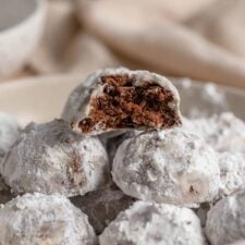 A bowl of icing sugar snowball cookies. The cookie on top has been bitten into so you can see the chocolate cookie with chocolate chips inside.