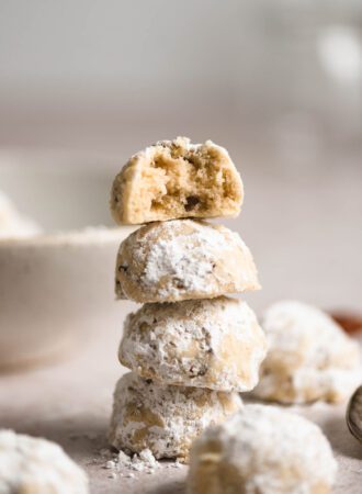 A stack of 4 pecan snowballs cookies coated in icing sugar. The one on top has a bite taken from it and you can see bits of pecans inside the dough.