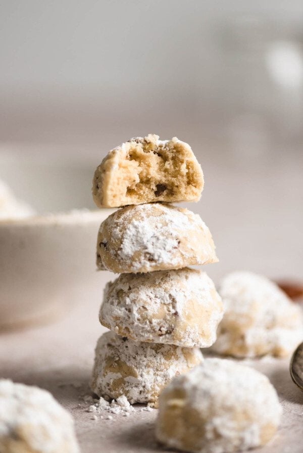 A stack of 4 pecan snowballs cookies coated in icing sugar. The one on top has a bite taken from it and you can see bits of pecans inside the dough.