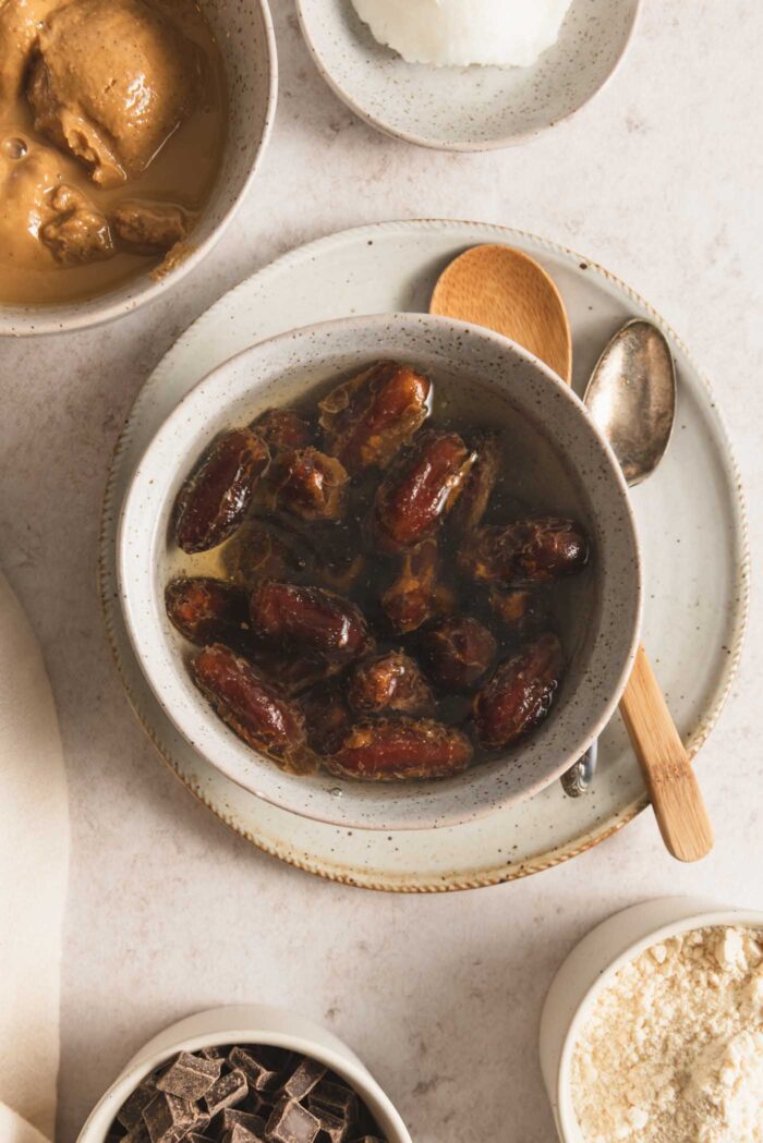 A bowl of dates soaking in water sitting on a plate with two small spoons resting on the plate.