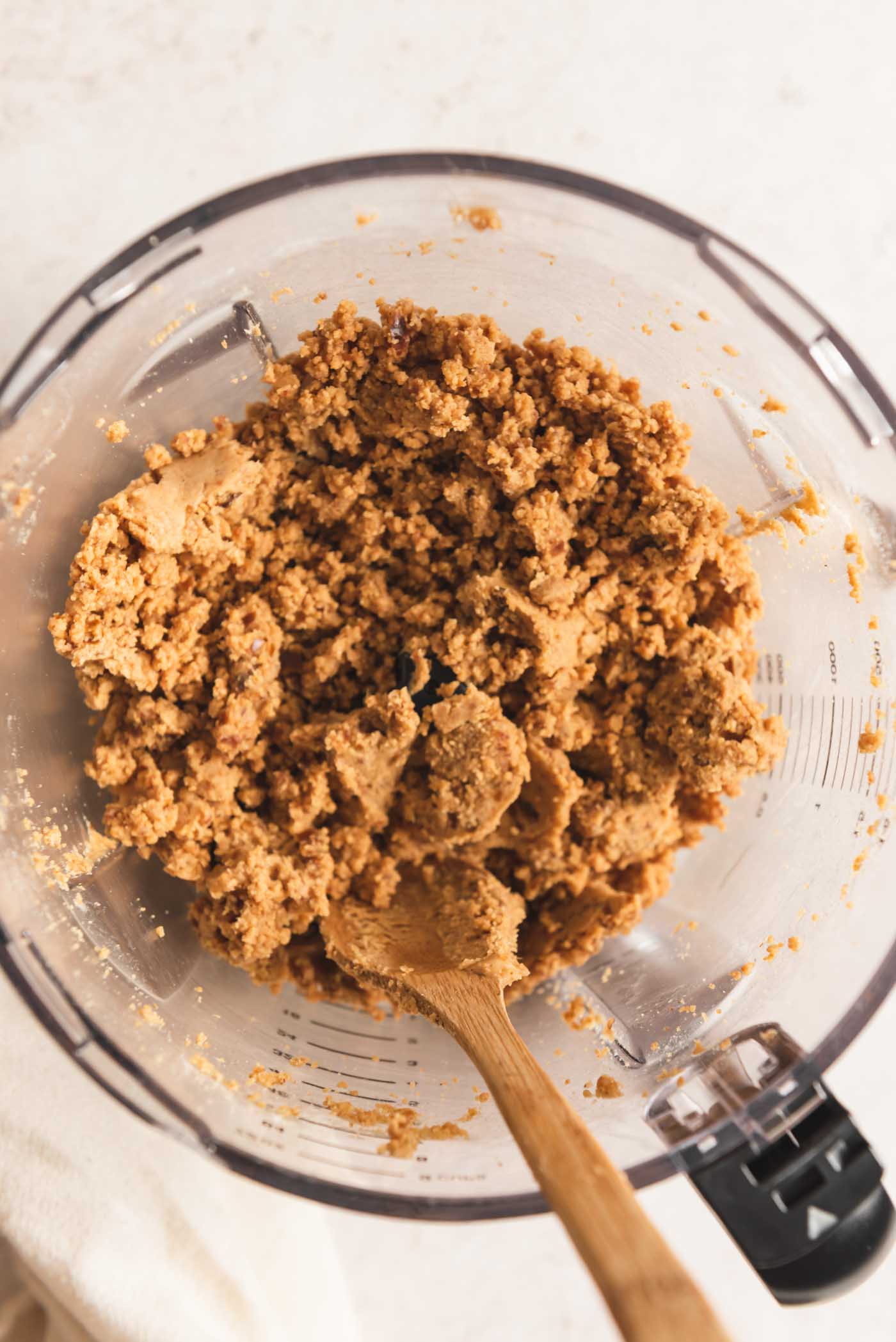 A crumbly peanut butter dough mixture in the container of a food processor with a small wooden spoon resting in it.