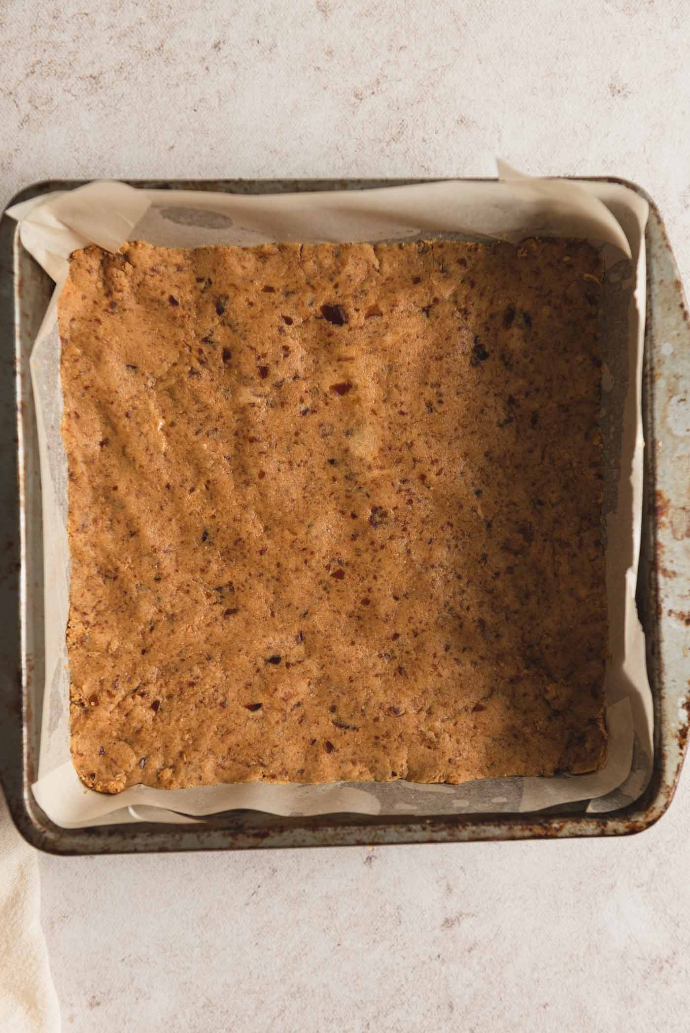 A layer of peanut butter dough for making peanut butter bars pressed into a square pan lined with parchment paper.