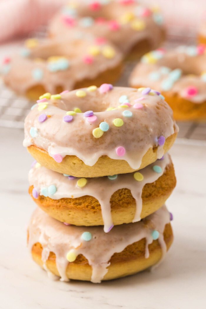 A stack of 3 glazed donuts with sprinkles with more donuts in the background.