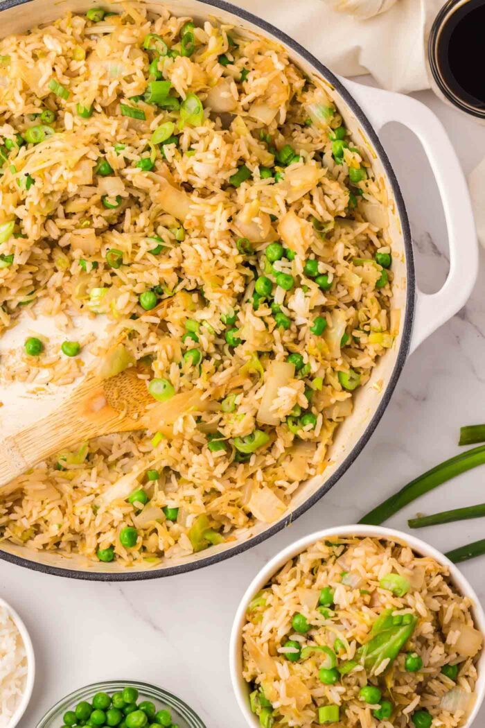A bowl of cabbage fried rice beside a large skillet of fried rice with a wooden spatula in it.