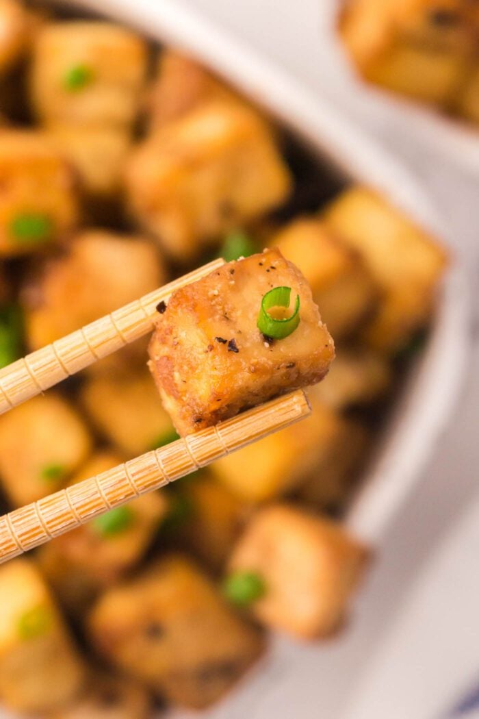 A pair of chop sticks holding a small cube of crispy tofu over a bowl of baked tofu.