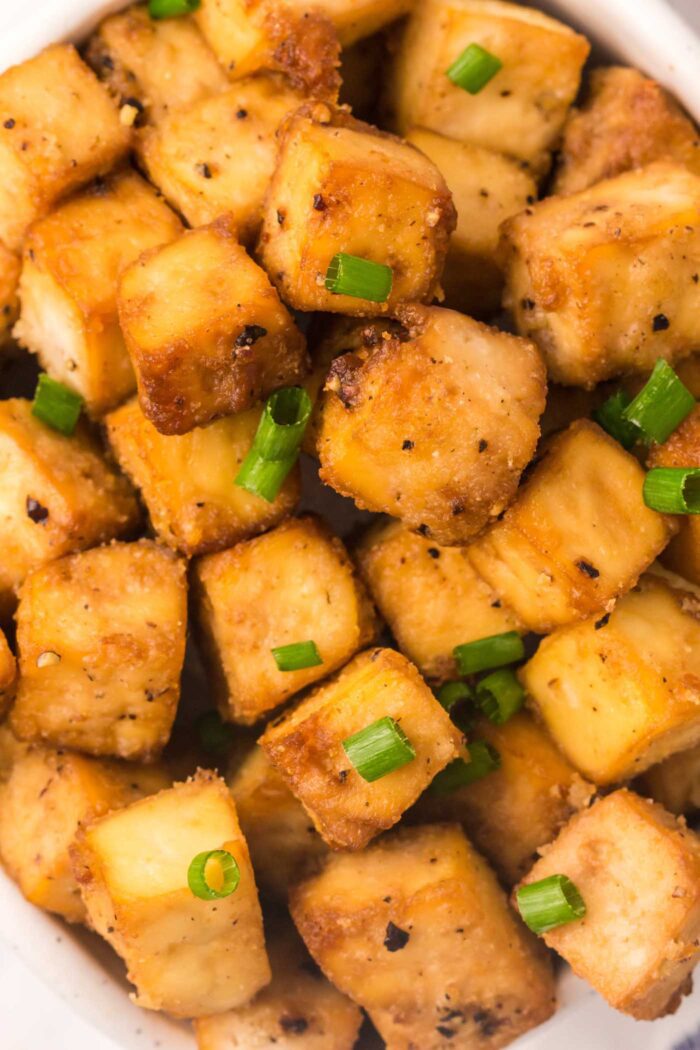 Close up of a bowl of crispy cubes of baked tofu with green onion sprinkled on them.