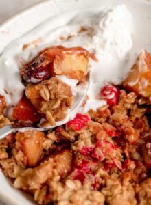 A bowl of cranberry and apple crisp topped with whipped cream with a spoon resting in the bowl.