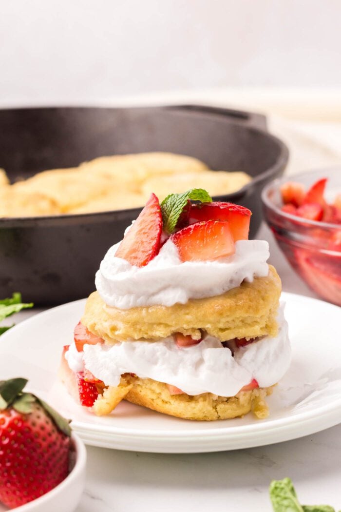A strawberry shortcake with a biscuit layered with whipped cream and strawberries on a small plate in front of a cast iron skillet of biscuits.