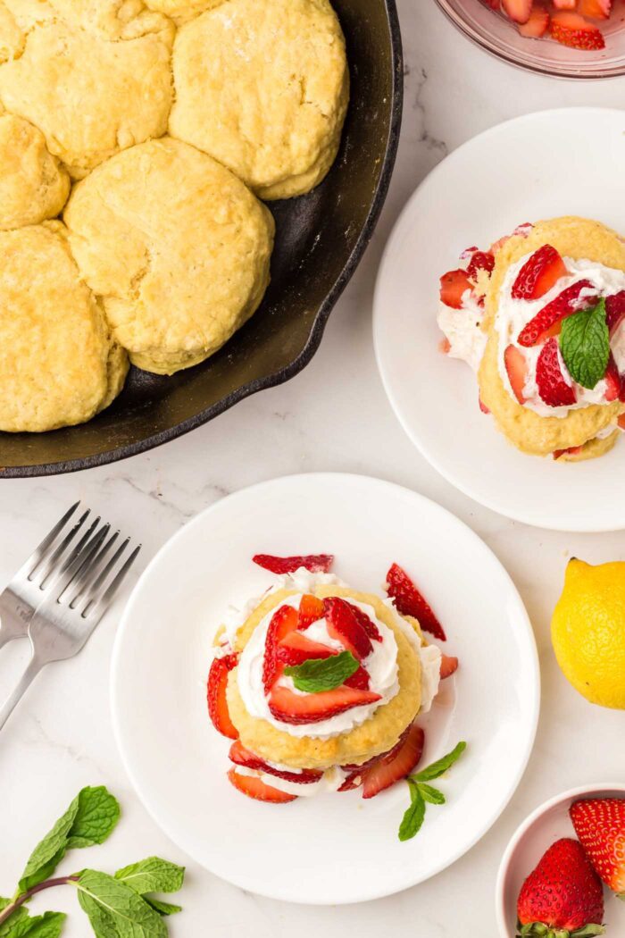Overhead view of two strawberry shortcakes topped with whipped cream and strawberries on small plate beside skillet of biscuits.