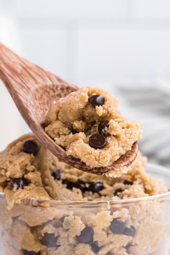 Edible chocolate chip cookie dough in a small glass bowl being scooped out with a small wooden spoon.