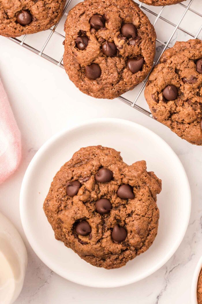 Overhead view of a double chocolate cookie with chocolate chips on top on a plate beside a cooling rack of more cookies.