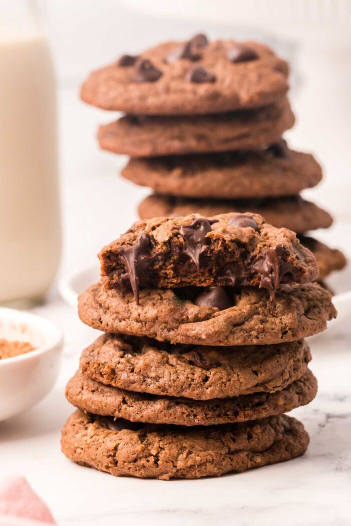 A stack of a number of double chocolate cookies with chocolate chips. The cookie on top has been broken in half so you can see the texture inside and some melted chocolate chips.