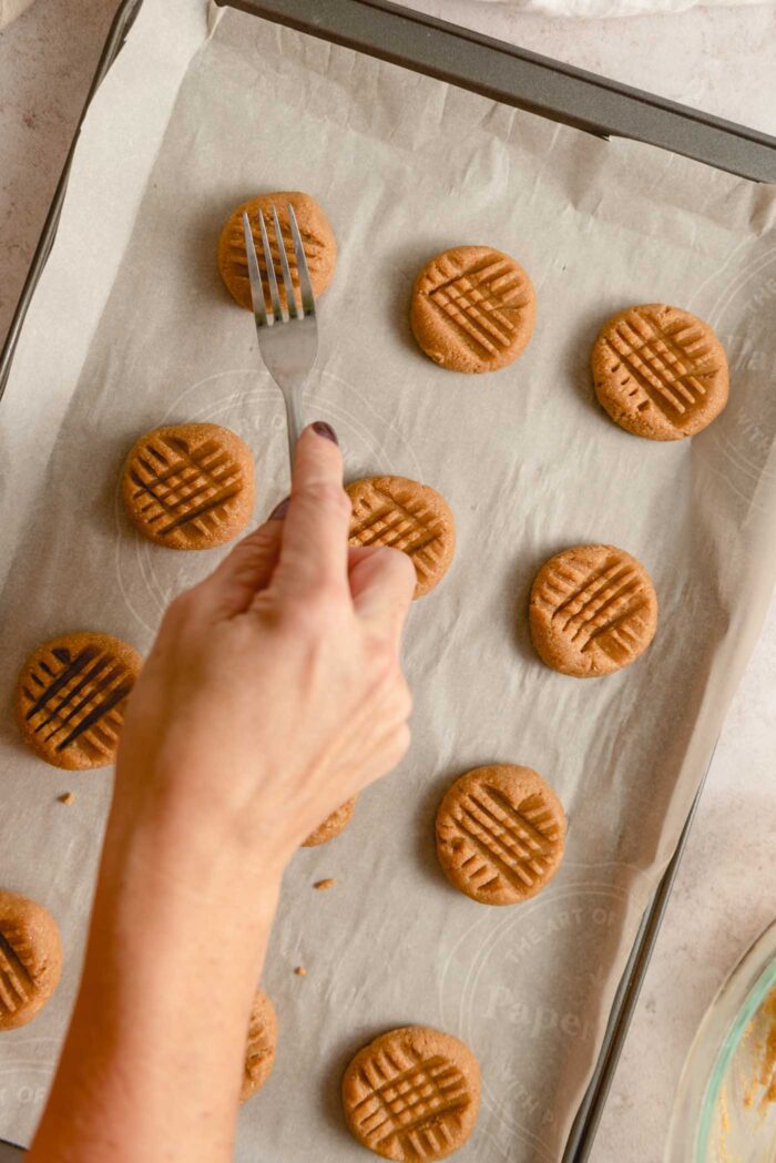 A hand using a fork to press a crisscross pattern onto peanut butter cookies on a baking tray.
