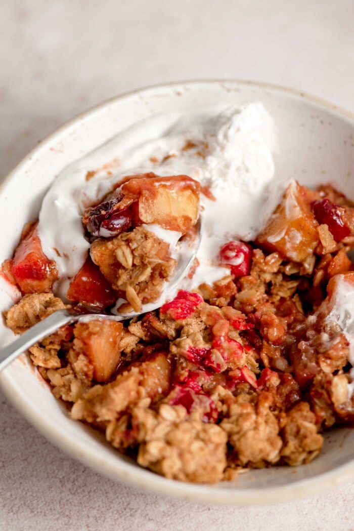 A bowl of cranberry apple crumble topped with whipped cream with a spoon resting in the bowl.