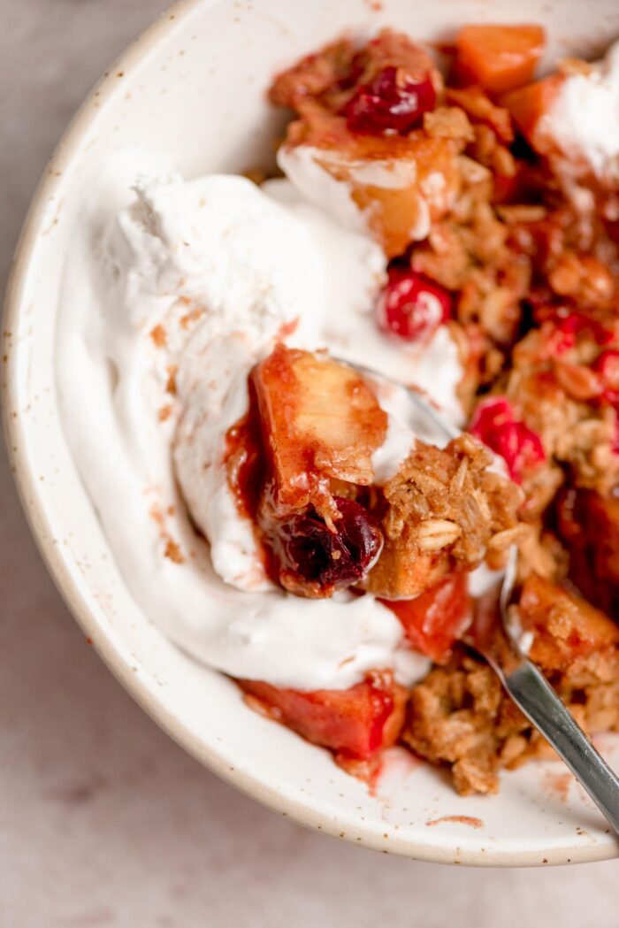 Close up of a bowl of cranberry apple crisp with whipped cream on top and a spoon in the bowl.