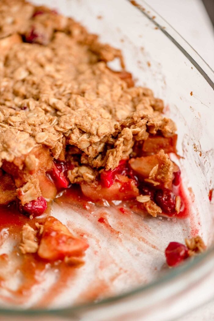 Close up of an apple cranberry crisp with a scoop out of it so you can see the apple cranberry filling and oatmeal crumble topping in a baking dish.