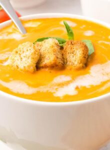 A bowl of creamy tomato soup drizzled with cream and topped with croutons and a piece of fresh basil.