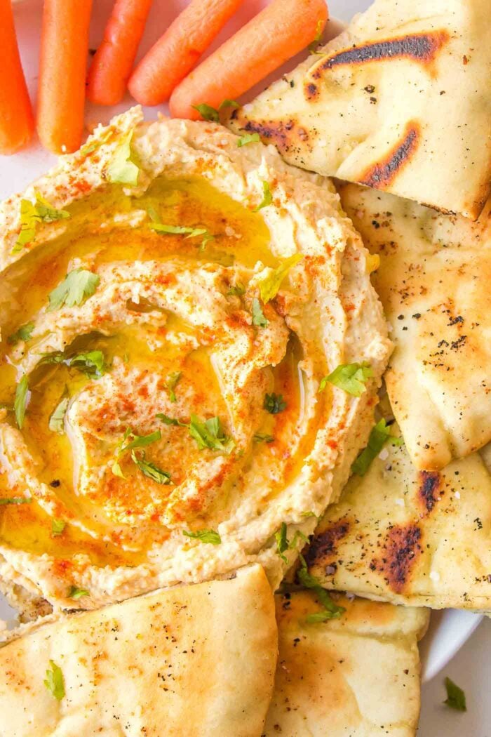A bowl of creamy roasted garlic hummus topped with olive oil and cayenne pepper with piece of pita and baby carrots around it.