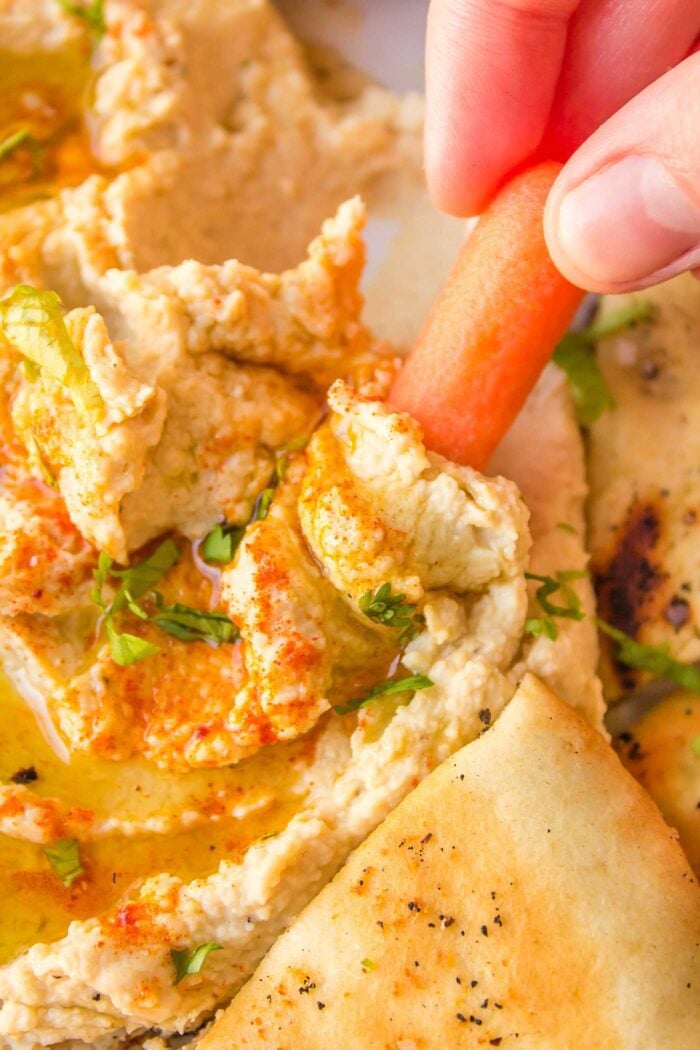 A carrot being dipped into hummus drizzled with olive oil.