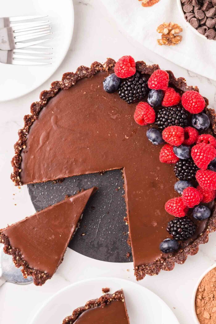 A chocolate ganache tart topped with fresh raspberries and blueberries with a slice out of it.