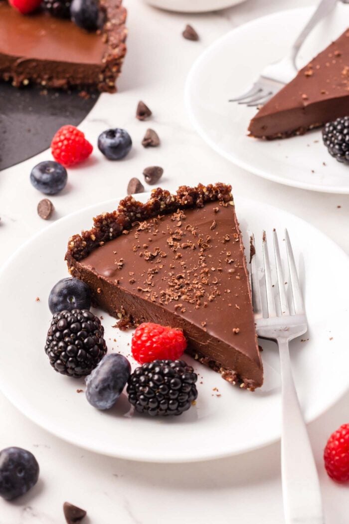 A slice of a chocolate tart with chocolate crust on a small plate with a few raspberries and blackberries on it.