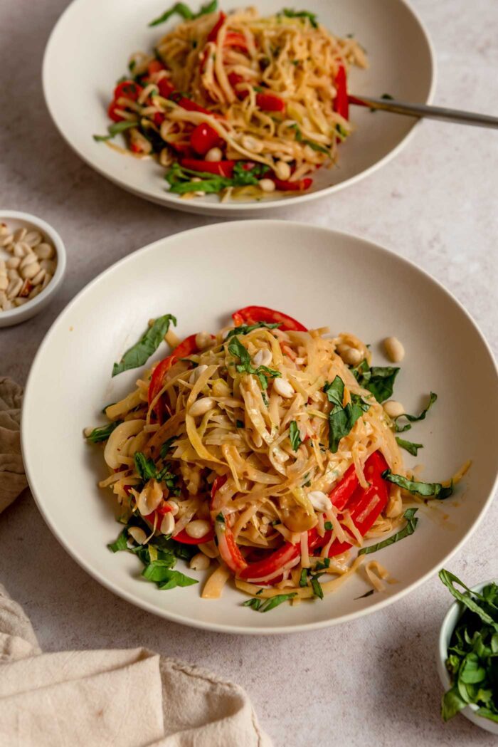 Two bowls of Thai red curry noodles with bell peppers and cabbage, topped with basil and peanuts.