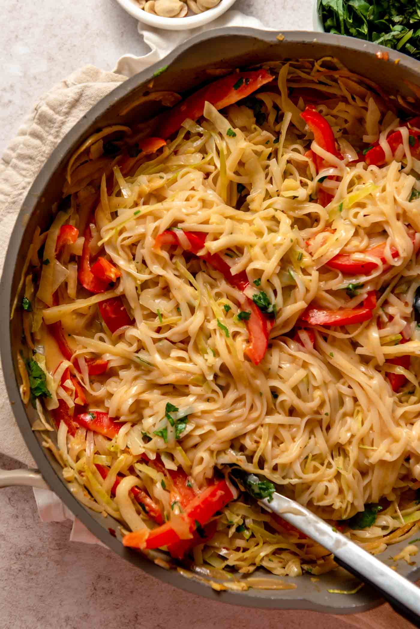 Overhead view of a large pan of Thai red curry noodles with cabbage and red pepper. A pair of tongs rests in the pan.