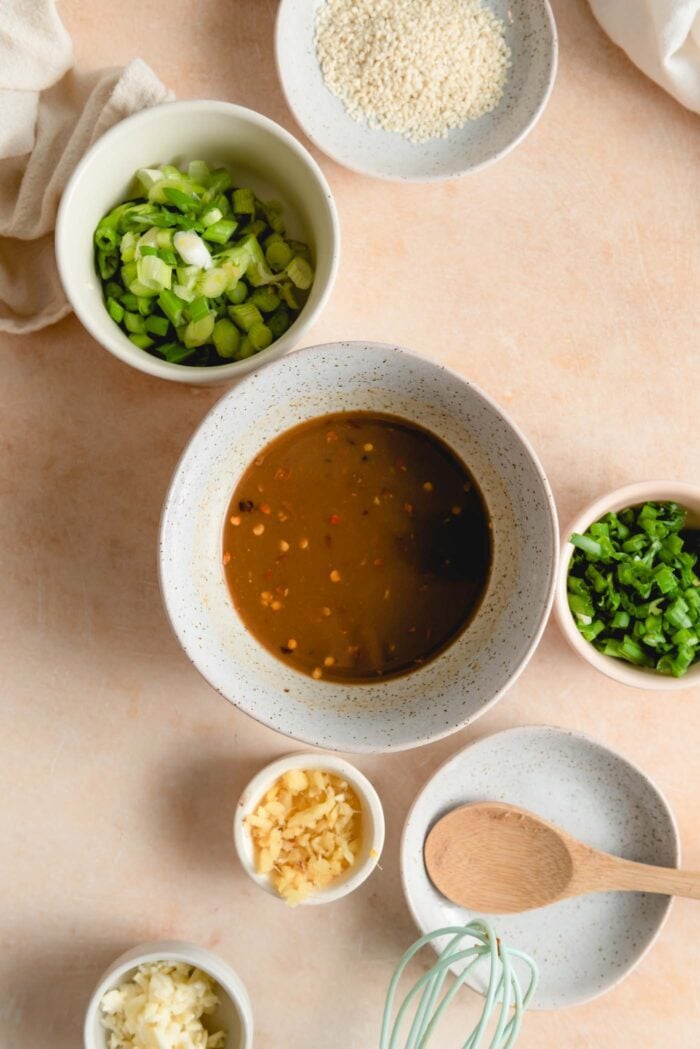A soy-based sauce with chili flakes in it in a small bowl with smaller bowls of minced garlic, ginger and green onion beside it.