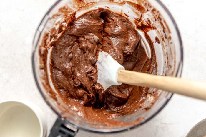 Chocolate brownie batter in a food processor container with a spatula resting in it.