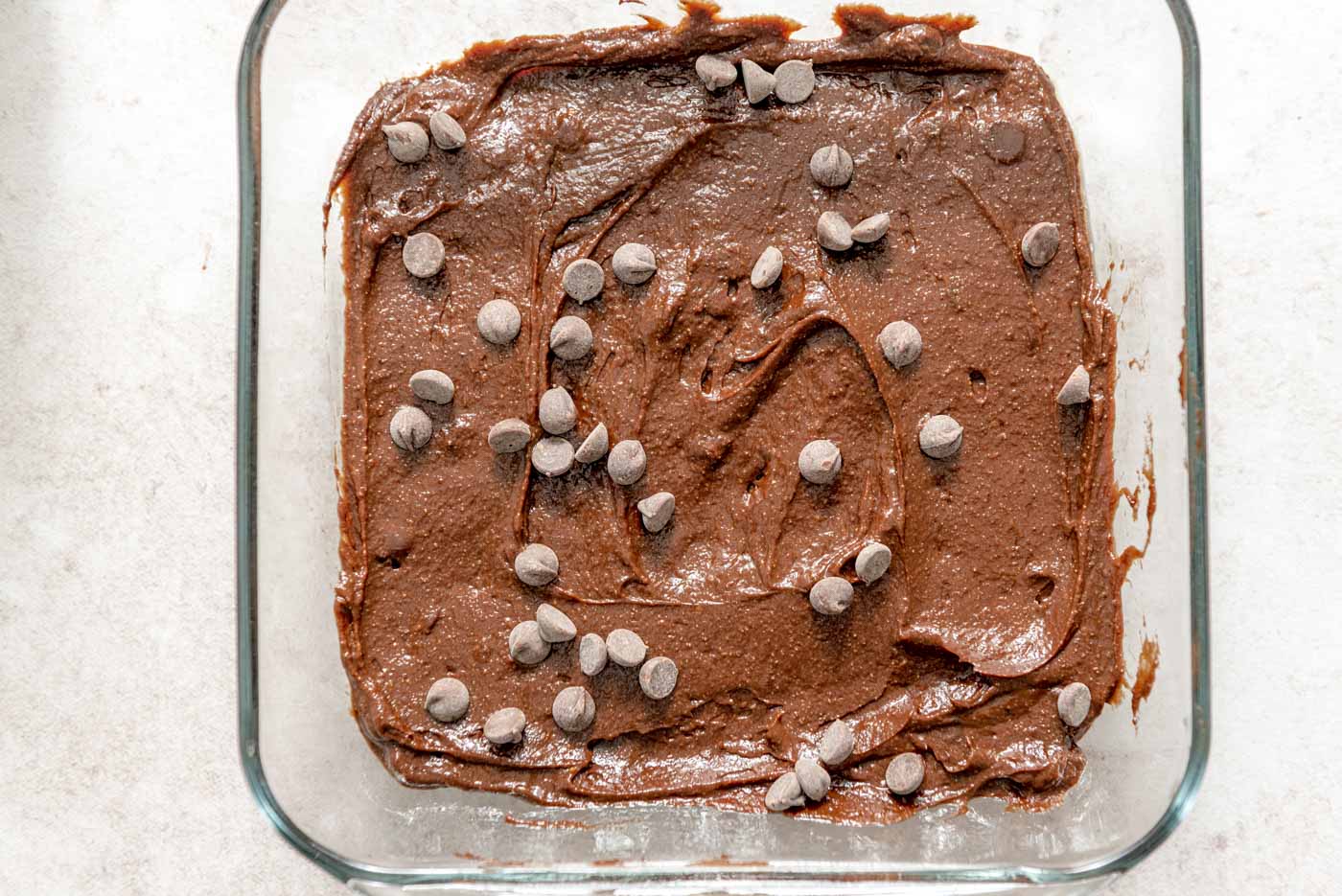 Raw brownie batter topped with chocolate chips in a glass baking dish.