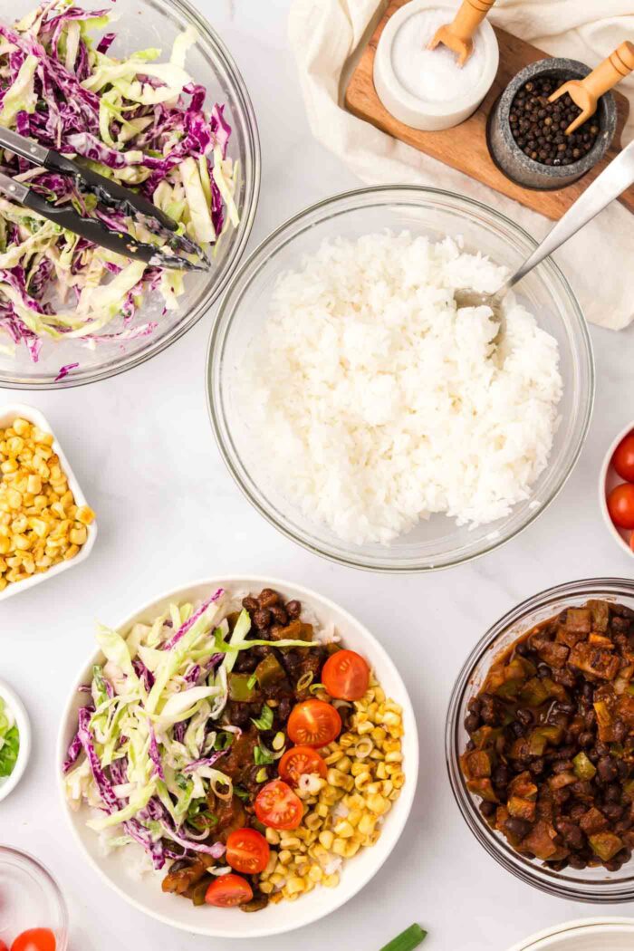 A bowl of rice with a spoon in it beside a bowl of cabbage slaw and a bowl with corn, tomato, bbq black beans and cabbage arranged in it.