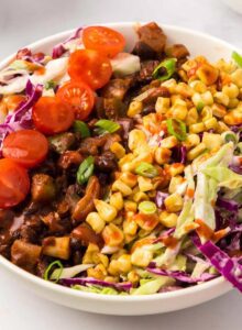 A colourful vegetable bean and rice bowl with corn, tomato and red and green cabbage slaw with BBQ sauce drizzled on top.