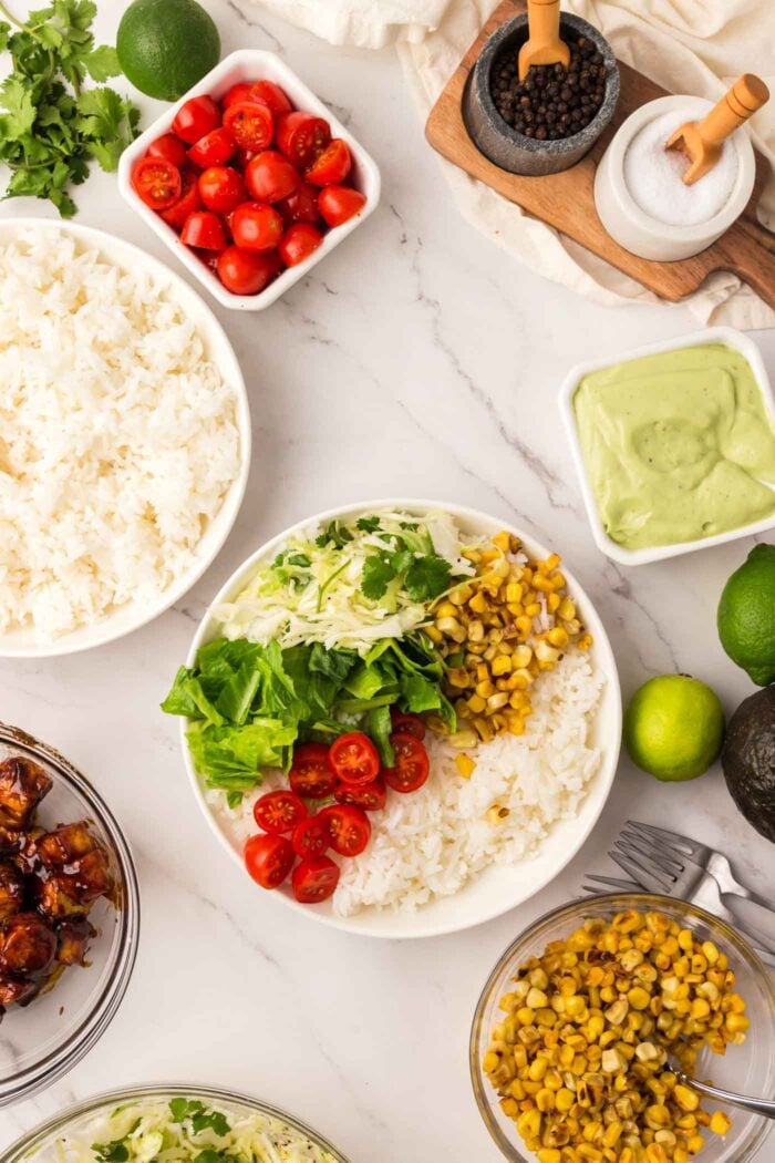 A bowl with rice, corn, lettuce, cabbage and grape tomatoes. There are bowls of corn, tomatoes, green salad dressing and rice beside it.