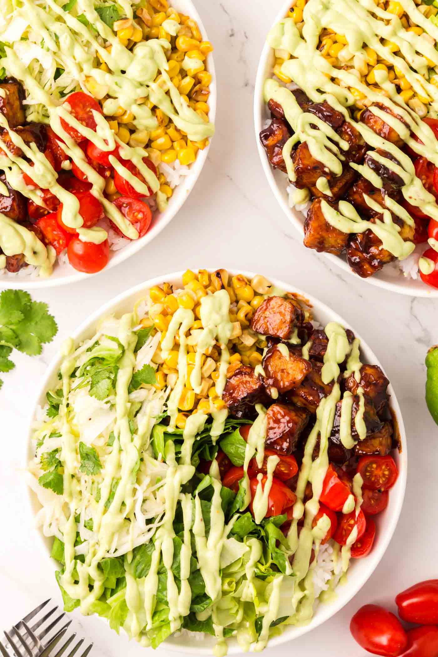 Above is an overhead view of three BBQ tempeh bowls with lettuce, corn, tomato, rice and avocado dressing.