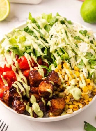 A BBQ tempeh bowl with corn, tomato, lettuce, and rice, drizzled with avocado dressing.