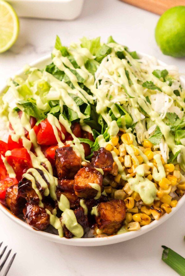 A BBQ tempeh bowl with corn, tomato, lettuce, and rice, drizzled with avocado dressing.