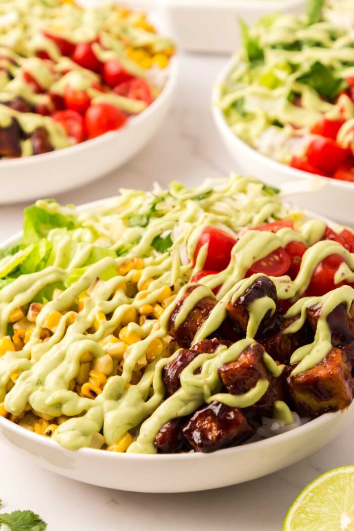 A BBQ tempeh bowl with corn, tomato, lettuce and cabbage topped with a drizzle of creamy avocado dressing.