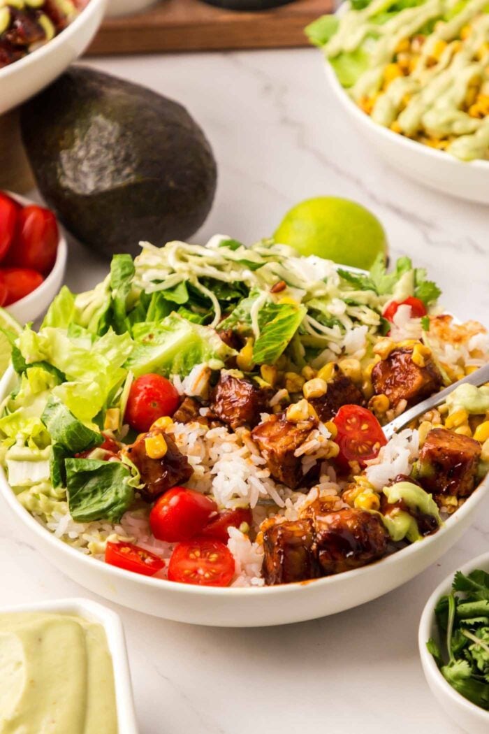 A BBQ tempeh bowl with lettuce, cabbage, corn and tomato. There is a small dish of dressing, an avocado, a small dish of cilantro and two more BBQ tempeh bowls in the scene.