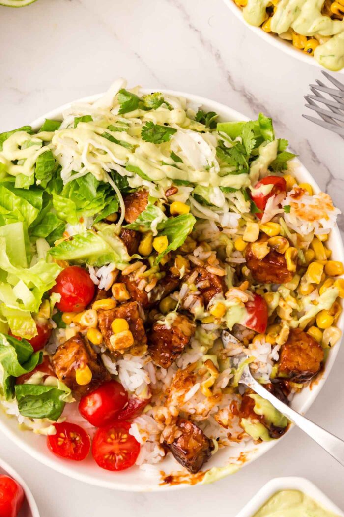 A mixed up BBQ tempeh bowl with corn, cabbage, lettuce, tomato and avocado dressing.