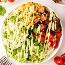 Overhead view of a BBQ tempeh bowls with lettuce, corn, tomato, rice and avocado dressing drizzled over top.