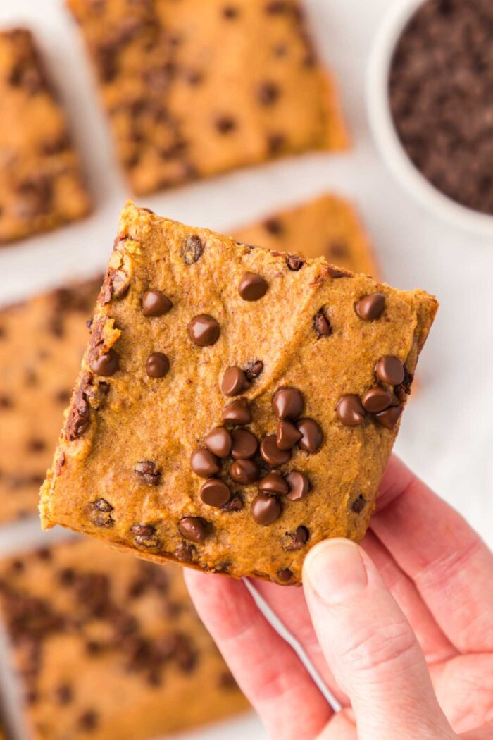 A hand holding a square of pumpkin chocolate chip bar over more bars on a countertop.